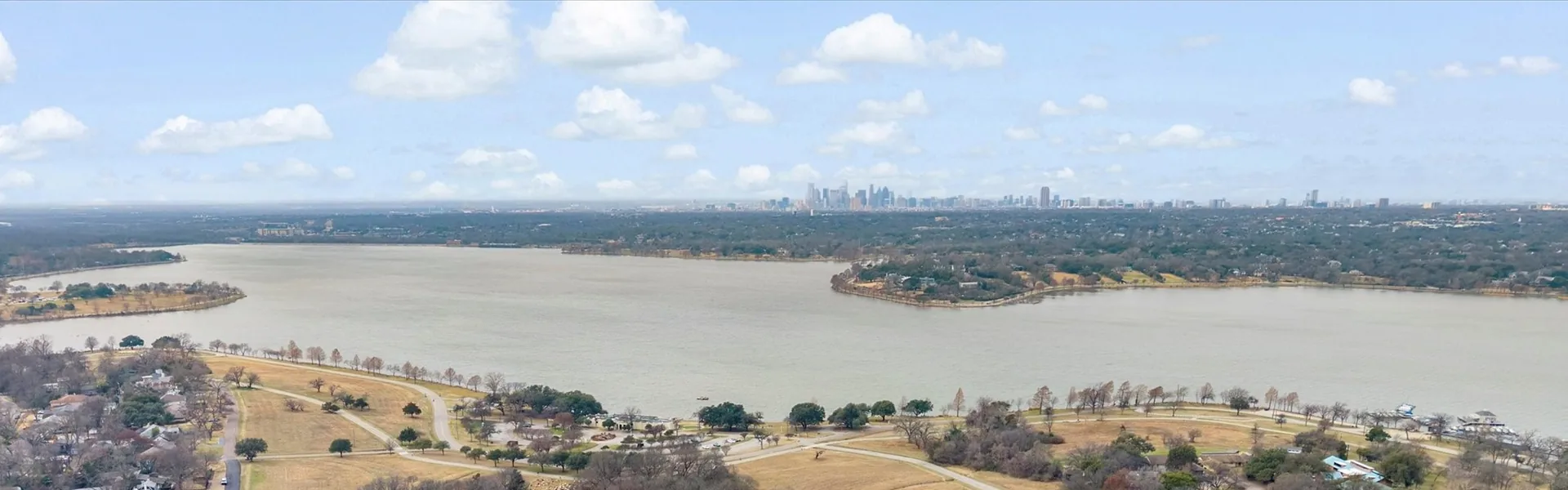 An aerial view over the residential streets of East Dallas towards White Rock Lake, with the downtown skyline on the horizon.